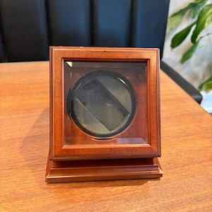May include: A wooden watch winder with a clear front panel. The winder has a square shape with a dark circular interior. The wood has a reddish-brown finish, and the base is rectangular. The winder is designed to keep automatic watches running.