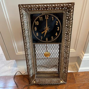 May include: Vintage rectangular wall clock with a black clock face and gold hands and numbers. The clock is encased in a decorative metal frame with an openwork design and a pendulum. The clock is plugged in.