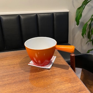 Pode incluir: Pote de fondue de cerâmica laranja e branca com alça, sobre um descanso de copo com padrão vermelho e branco em uma mesa de madeira.