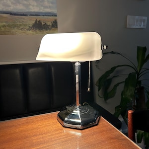 May include: A chrome-finished banker's lamp with a white glass shade, resting on a wooden table. The lamp features a pull chain and a geometric base. The background includes a painting and a plant.