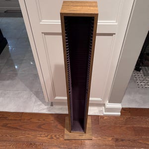 May include: A tall, vertical media storage tower with a wood grain finish. The unit features a narrow, open-front design with multiple horizontal slots along the interior sides for organizing discs or small media cases, supported by a square base.
