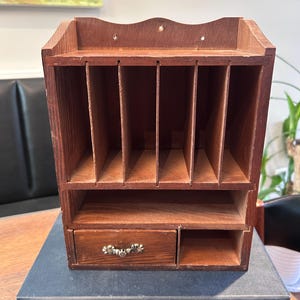 May include: A brown wooden desk organizer featuring five vertical slots at the top, a horizontal shelf below, and a small drawer with a decorative metal handle next to an open compartment. The organizer rests on a dark blue book titled Whom Fortune Favours.