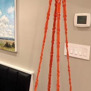 May include: Orange macrame plant hanger with a clear, round base. The hanger is suspended from a knotted top and features four twisted rope strands. Decorative orange tassels are attached to the base. A stylish way to display plants.