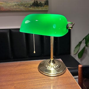 May include: A banker's lamp featuring a vibrant green glass shade and a polished brass base. The lamp includes a vertical fluted stem, a circular tiered pedestal, and a gold-toned pull chain with a small finial for turning the light on and off.