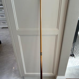 May include: A wooden pool cue with a light brown shaft and a dark brown butt. The cue has a black band near the middle and a white tip. The cue is standing upright against a white cabinet.