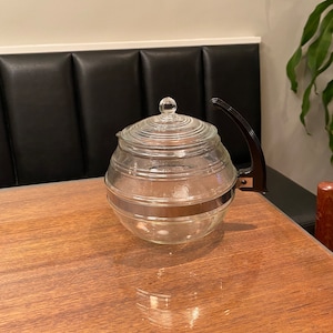 Puede incluir: Una tetera de vidrio transparente con cuerpo redondo y tapa con un pequeño pomo. La tetera tiene un asa negra y una banda plateada en el centro. Se encuentra sobre una mesa de madera.