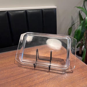 May include: A clear rectangular glass lid for a baking dish with integrated side handles. The word Pyrex is embossed on the left side of the rim, and additional text is embossed on the right side of the rim. The lid is held upright by a black stand.