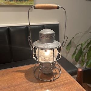 Könnte beinhalten: Vintage Metalllaterne mit klarer Glaskugel und Holzgriff. Die Laterne hat ein käfigartiges Design und wird von innen beleuchtet, wodurch ein warmes Leuchten entsteht. Die Laterne steht auf einem Holztisch.