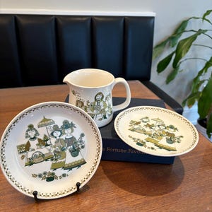 May include: A vintage cream-colored ceramic pitcher and two plates with a green and brown illustration of women in dresses and hats. The plates are round and oval, with a decorative border. The pitcher has a handle and a similar design.