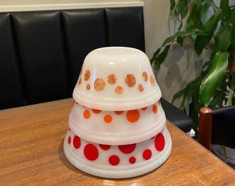 May include: Three white mixing bowls with red and orange polka dots stacked on top of each other. The bowls are on a wooden table.