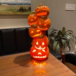 May include: A tower of six illuminated orange ceramic jack-o'-lanterns with carved faces. The pumpkins are stacked on top of each other, creating a Halloween decoration. The pumpkins are sitting on a wooden table. The warm light creates a spooky atmosphere.