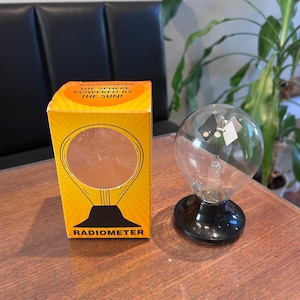 May include: A glass radiometer with a black base and internal rotating vanes, accompanied by a yellow rectangular box. The box features the text RADIOMETER THE SPHERE POWERED BY THE SUN! and a graphic illustration of the device on the front.