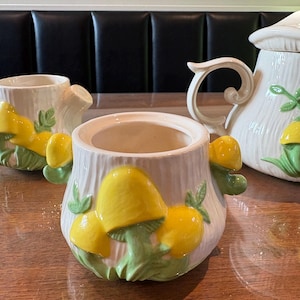 May include: A white ceramic sugar bowl with a tree trunk design, decorated with yellow mushroom accents and green leaves. The bowl is part of a tea set, with a teapot and creamer visible in the background.