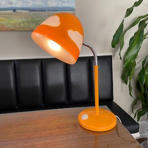 May include: Orange and white mushroom-shaped desk lamp with a flexible arm. The lamp is on a wooden table.