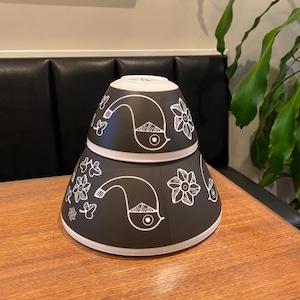Puede incluir: Dos cuencos de cerámica negra con diseños florales y de pájaros blancos apilados uno encima del otro. Los cuencos están sobre una mesa de madera oscura.