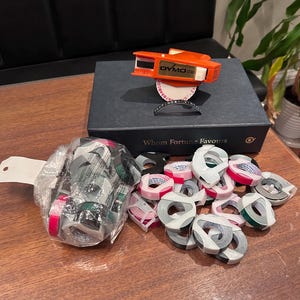 May include: An orange Dymo 1780 embossing label maker sits atop a black box labeled Whom Fortune Favours. Beside the box is a clear plastic bag filled with multiple rolls of colored embossing tape, along with several loose rolls in pink and grey.