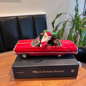 Könnte beinhalten: Ein rotes Spielzeug-Cabriolet mit dem Weihnachtsmann auf dem Fahrersitz und zwei Rentieren mit grünen Mützen als Passagiere. Das Auto steht auf einer schwarzen rechteckigen Schachtel mit dem goldenen Aufdruck "MUSSIO" und "Whom Fortune Favours".
