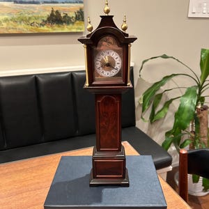 Puede incluir: Un reloj de pie en miniatura con acabado en madera oscura, detalles dorados y una esfera con números romanos. El reloj se exhibe sobre un libro azul oscuro con el texto "Whom Fortune Favours".
