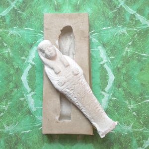 May include: An off-white sarcophagus figurine with hieroglyphs and a detailed face, in a rectangular mold. The background is a green and white patterned surface. The figurine is a representation of ancient Egyptian art.