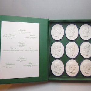 Könnte beinhalten: Eine grüne Box mit weißem Innenraum, die 10 weiße Keramikmedaillons mit dem Profil römischer Kaiser enthält. Die Medaillons sind in drei Reihen angeordnet. Der Deckel der Box hat einen weißen Papiereinsatz mit den Namen der Kaiser und ihren Regierungszeiten.
