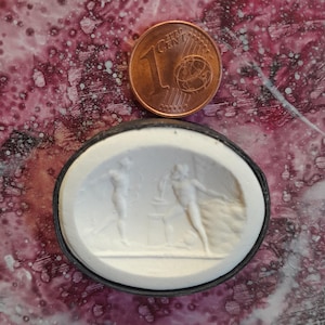 Könnte beinhalten: Ein weißer Kamee mit schwarzem Rand, der zwei Figuren im klassischen Stil darstellt. Der Kamee ist etwa so groß wie eine deutsche Ein-Cent-Münze.