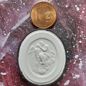 Puede incluir: Un camafeo ovalado blanco con un diseño en relieve de una figura alada que es sostenida por una figura más grande. El camafeo está engastado en un marco ovalado negro.