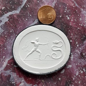 Op de afbeelding: Een witte gipsen reliëfsculptuur van een figuur met een boog en pijl, die tegenover een slang staat. De sculptuur is rond en heeft een zwarte rand. De tekst "DIANA" is zichtbaar aan de onderkant van de sculptuur.