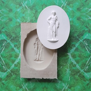 Pode incluir: Uma escultura em relevo ovalada branca de uma figura masculina nua, em pé com um pano drapeado. A escultura está ao lado de um molde da mesma figura. O fundo é uma superfície com padrão verde e branco.