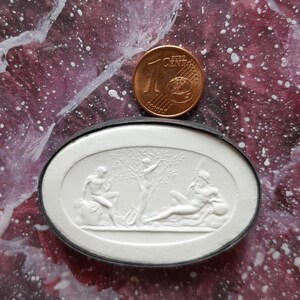 Könnte beinhalten: Ein weißer ovaler Kamee mit einem erhabenen Design aus drei Figuren. Die Figuren sind ein Mann, der in einem Baum steht, ein Mann, der auf einem Felsen sitzt, und ein Mann, der sich auf einem Felsen ausruht. Der Kamee ist in einen schwarzen ovalen Rahmen gefasst.