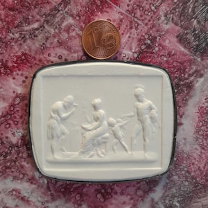 Puede incluir: Una escultura en relieve rectangular blanca con un borde negro, que representa figuras clásicas. Una moneda de un céntimo de euro se coloca encima de la escultura. La moneda tiene aproximadamente 1,9 cm de diámetro.