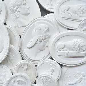 Könnte beinhalten: Eine Sammlung weißer Gipsreliefplatten mit klassischen Figuren und Tieren. Die Platten variieren in Form und Größe und zeigen detaillierte Darstellungen von Büsten, Löwen und mythologischen Szenen. Die Gesamtästhetik ist antik und künstlerisch.