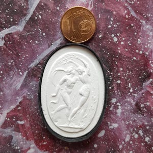 Könnte beinhalten: Ein weißer Gips-Kamee mit einem Relief von zwei tanzenden Figuren. Der Kamee ist in einem schwarzen ovalen Rahmen gefasst. Eine 1-Euro-Münze dient als Größenvergleich.