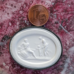 Könnte beinhalten: Ein weißer ovaler Kamee mit einem erhabenen Design aus drei Figuren. Die Figuren sind ein laufender Mann, eine Frau, die ein Kind hält, und ein Kind, das die Hand zur Frau ausstreckt. Der Kamee ist in einem schwarzen ovalen Rahmen gefasst.