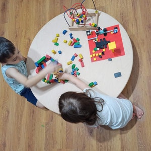 Peut inclure: Deux enfants jouent avec des blocs de bois colorés et un ensemble Lego rouge et noir sur une table ronde en bois.