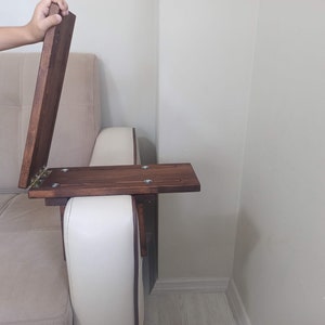 May include: A wooden table with a hinged top that can be folded down. The table is attached to the arm of a sofa.