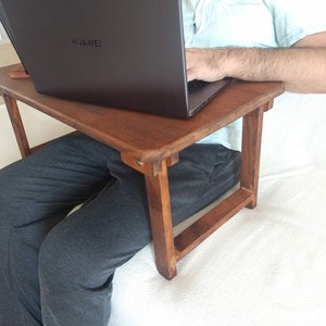 May include: A small, rectangular, wooden laptop desk with four legs. The desk is a warm brown color and is being used with a laptop. The laptop has the brand name "HUAWEI" on the top. The desk is designed to be used while sitting.