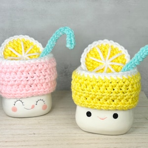 May include: Two white ceramic mugs with cute faces, each topped with a crocheted lemon slice and a blue crocheted straw. The mug on the left has a pink crocheted base, while the mug on the right has a yellow crocheted base.