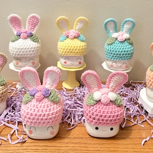 May include: Seven ceramic mugs with bunny ears and crocheted hats in various colors, including pink, blue, yellow, and white. Each mug has a smiling face and is decorated with flowers.