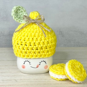 May include: A white ceramic mug with a smiling face, topped with a yellow crocheted lemon-shaped cozy and a green leaf. Two yellow and white crocheted macarons sit beside the mug. The cozy is tied with twine.