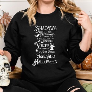 K&ouml;nnte beinhalten: Ein schwarzes Sweatshirt mit einem wei&szlig;en Grafikdruck, der "Shadows of a thousand years rise again unseen voices whisper in the trees tonight is Halloween" lautet.