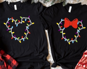 Puede incluir: Dos camisetas negras con un diseño de luces navideñas en forma de Mickey Mouse y Minnie Mouse. La camiseta de Mickey Mouse tiene un diseño simple, mientras que la camiseta de Minnie Mouse tiene un lazo rojo.
