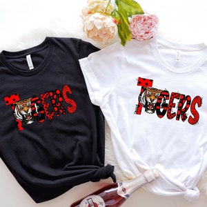 Könnte beinhalten: Zwei T-Shirts mit dem Wort "TIGERS" in roten und schwarzen Buchstaben mit einem Tigergesicht und Punkten. Die Shirts liegen auf einer weißen Oberfläche.