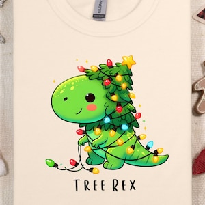 May include: A cream-coloured t-shirt featuring a cartoon illustration of a green dinosaur wearing Christmas lights and a star on top of its head. The text "TREE REX" is printed below the dinosaur.