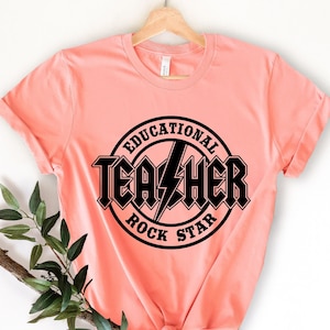 以下が含まれることがあります： ピーチ色のTシャツに、黒い円形のグラフィックがプリントされています。グラフィックには「Educational Rock Star」と書いてあり、単語「Teacher」の真ん中に稲妻が描かれています。