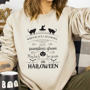 Peut inclure: Un sweat-shirt beige avec un imprim&eacute; graphique noir qui dit "When black cats prowl and pumpkins gleam, may luck be yours on Halloween". Le graphique repr&eacute;sente deux chats noirs, un chapeau de sorci&egrave;re et une citrouille.
