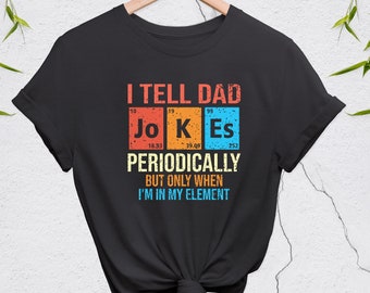Puede incluir: Camiseta negra con un diseño gráfico blanco y naranja. El gráfico presenta la palabra "JOKES" con las letras representadas como elementos químicos. El texto dice "Le digo chistes a papá periódicamente, pero solo cuando estoy en mi elemento."