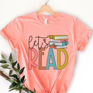 May include: A pink t-shirt with the text "let's read" in colorful letters. A stack of books with colorful covers is behind the text.