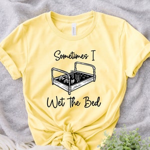 K&ouml;nnte beinhalten: Gelbes T-Shirt mit einer schwarz-wei&szlig;en Grafik eines Bettes mit darin wachsenden Pflanzen. Der Text "Sometimes I Wet the Bed" ist unter der Grafik aufgedruckt.