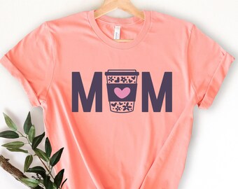 Pode incluir: Uma camiseta cor de pêssego com a palavra "MOM" impressa em letras pretas. Uma xícara de café com um coração rosa e um desenho floral está entre os "M".