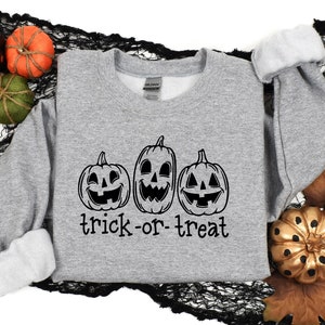 Może przedstawiać: Szara bluza z trzema uśmiechniętymi dyniami w czarnym tuszu. Pod dyniami wydrukowano napis "trick-or-treat". Rękawy są podwinięte, a tło zawiera dekoracyjne dynie i liście.